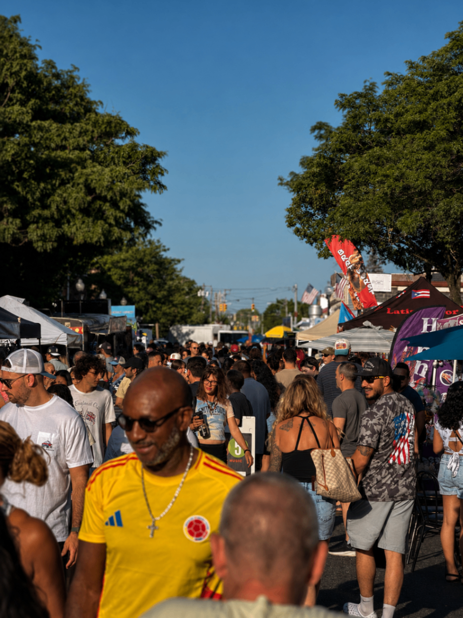 Packed summer street festival at golden hour with food tents, flags and a lively crowd walking the block