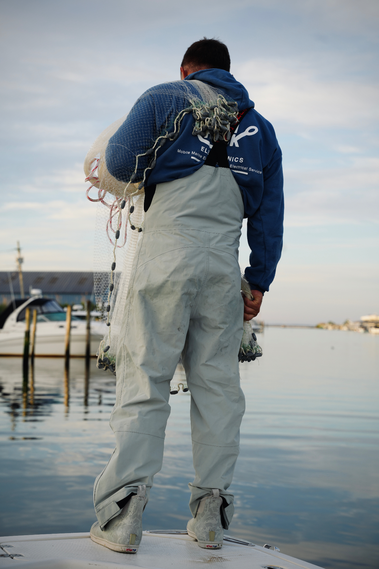 Fisherman in waders and hoodie casting a net from the bow of a boat at a calm marina