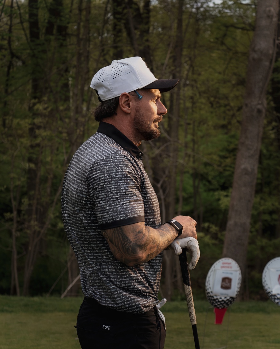 Tattooed golfer in a white cap and patterned polo resting on his club at a wooded tee box