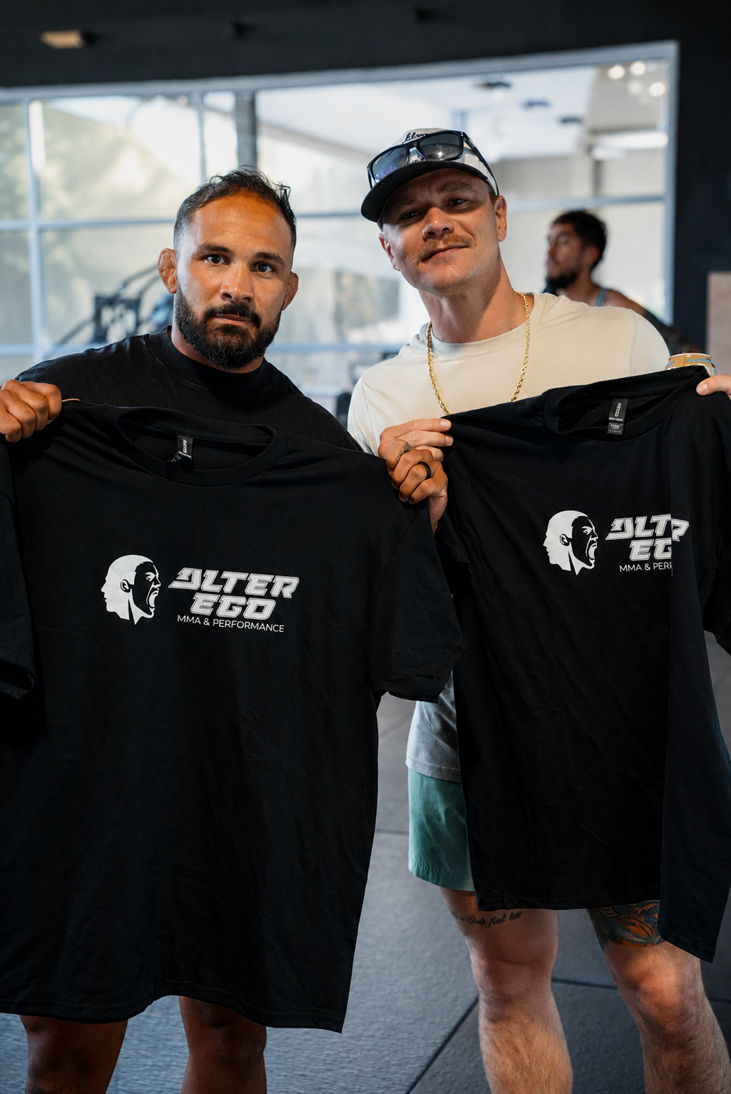 Two fighters holding up black Alter Ego MMA & Performance tees inside a training facility